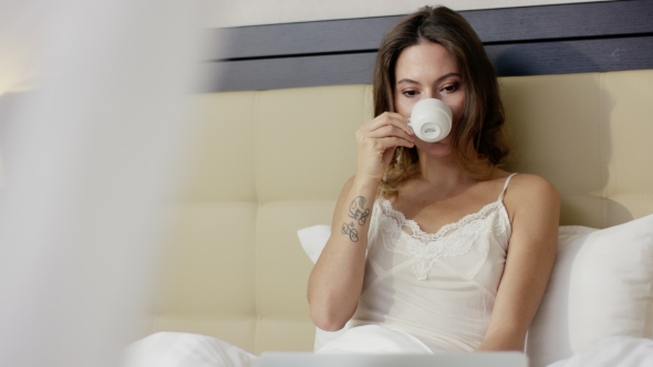 Young Woman Watching Movie on Laptop While Drinking Coffee in Bed