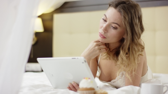 Attractive Woman Texting Over a Tablet Pc While Lying on Bed Near Cup of Coffee and Cupcake