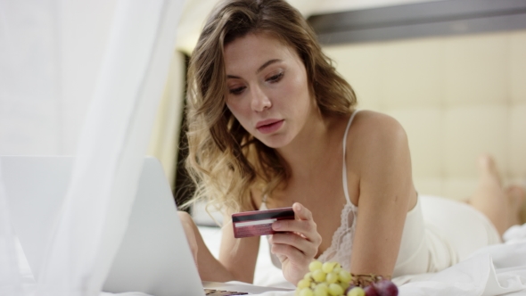 Pretty Woman Dressed in Sleepwear Lies on Bed and Using Her Laptop and Credit Card