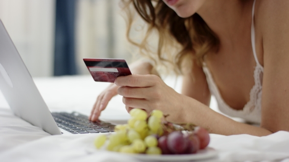 Charming Woman Dressed in Sleepwear Lies on Bed and Using Her Laptop and Credit Card alt