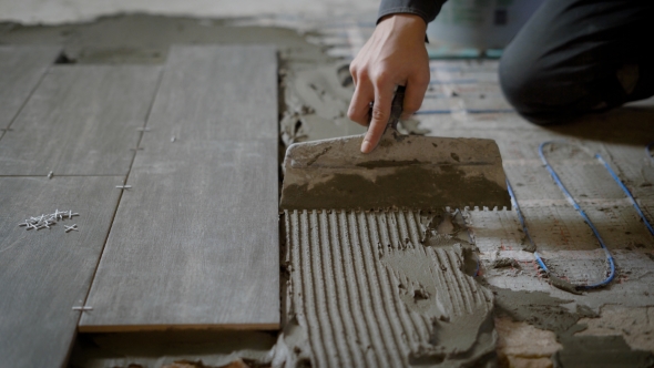 Masters Hand Is Holding Spatula with Teeth and Equalizing Building Mortar on a Floor During Process alt