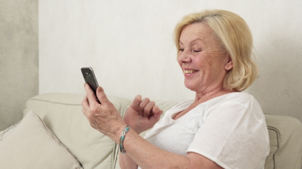 Happy Old Woman Sitting on Sofa and Using a Smart Phone