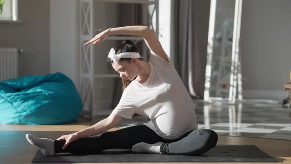 Flexible Young Pregnant Woman Bending Aside Sitting on Exercise Mat on Sunny Morning at Home alt