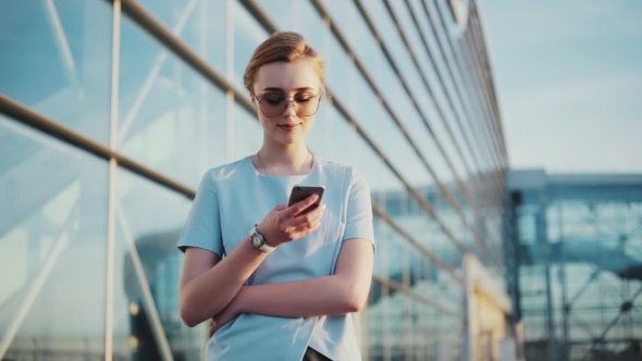Stylish Young Woman in Trendy Sunglasses Uses Her Mobile Phone, Outside the Glassy Urban Building on