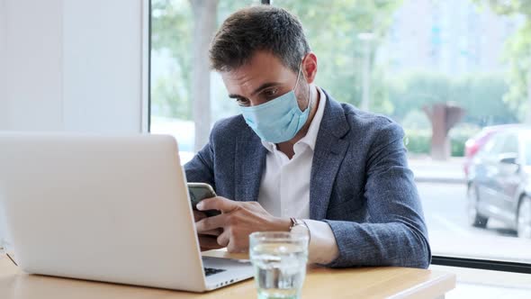 Businessman wearing protective mask using smartphone at laptop in cafe alt