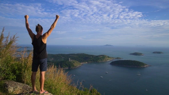 Athletic Young Man Stands with His Hands Up on the Edge of the Mountain. alt