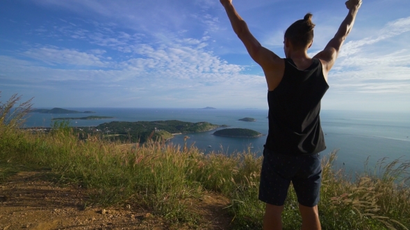 Athletic Young Man Stands with His Hands Up on the Edge of the Mountain. alt