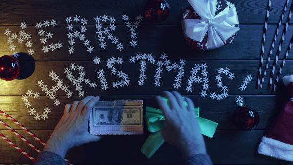 Adult Man Puts Cash Money Into New Year Gift Box on Wooden Decorated Table, Top Down Shot