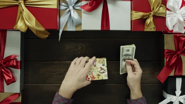 Adult Man Puts Dollars Into Xmas Present Box on Wooden Decorated Table, Top Down Shot