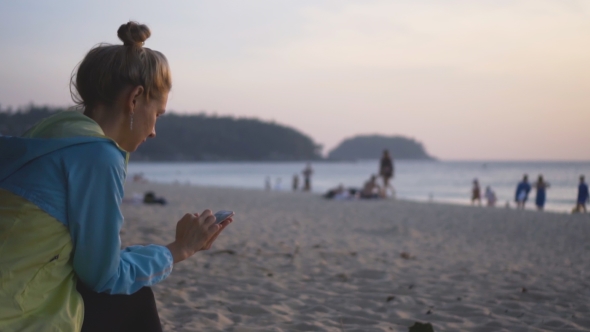 Sad Girl with a Phone Sits on the Beach at Sunset.