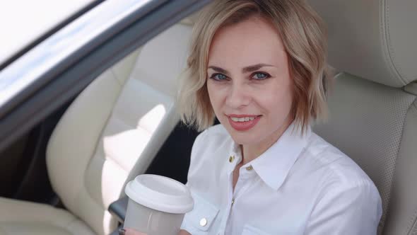 Smiling Business Woman in White Shirt Sitting in the Car and Drinking Coffee alt