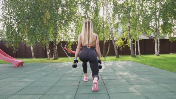 Muscular Woman Athlete Doing Walking Lunges with Dumbbells on a Sports Ground