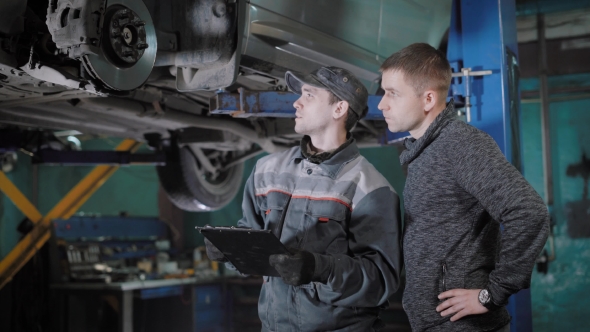 Automobile Mechanic Is Appraising a Brake Disk, Brake Pads and Hosepipe, Explaining for a Car Owner alt