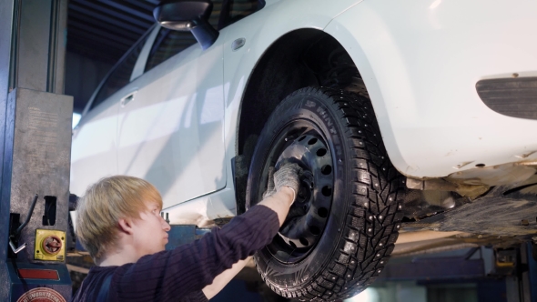 Auto Mechanic Is Twisting Manually Bolts of a Wheels on an Automobile, Raised By Lifting Equipment alt