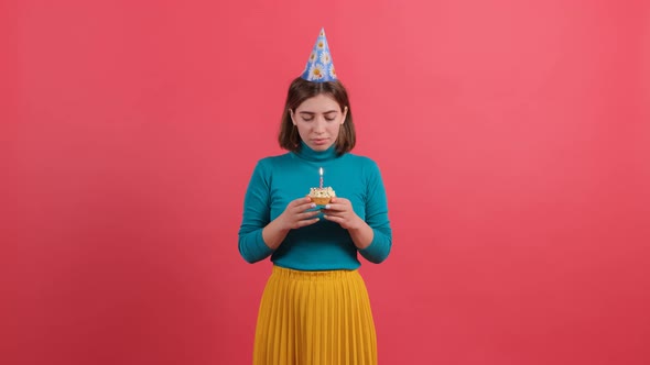 Young Woman in Birthday Hat Blowing Out Candle on Cake Isolated on Red Background alt
