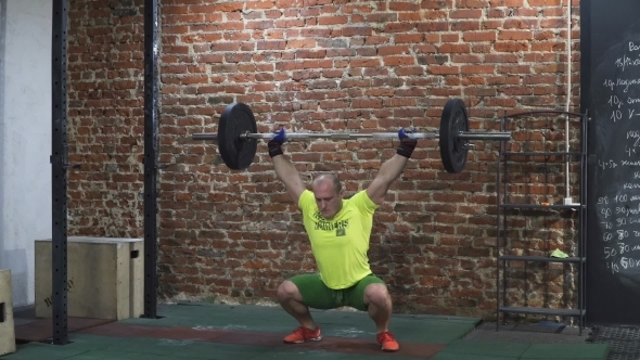 Man Athlete Doing Squats with a Barbell Over His Head in Cross Fit Gym