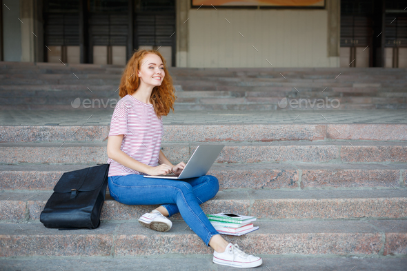 Smiling young redhead girl working with laptop sits on the stairs outdoors - Stock Photo - Images