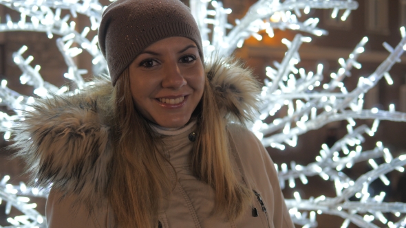 Young Woman in Warm Clothing Smiling Standing amid Christmas Illuminations alt