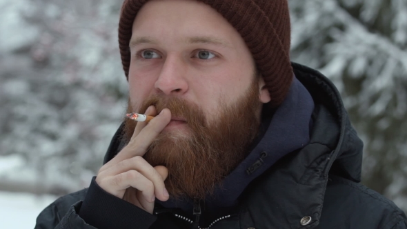 Young Bearded Hipster Smoking Cigarette in Hat and Jacket at Forest