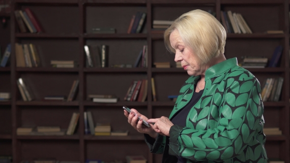Serious Elderly Woman Uses a Smartphone in the Library