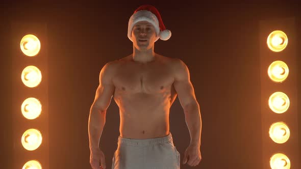 Portrait of Muscular Man Wearing Christmas Santa Hat Standing on Smoky Background alt