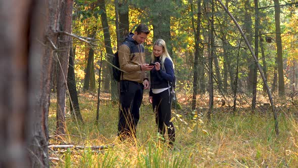 A Hiking Couple Stands in the Middle of a Meadow in a Forest and Looks at a Smartphone alt