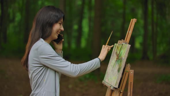 Middle Side View of Woman Artist Painting a Picture in the Park and Talking By the Phone alt