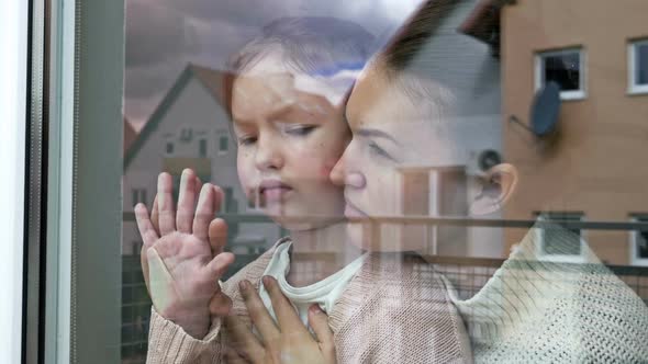 Mother and Little Daughter Stand Embracing and Look Sadly Out the Window