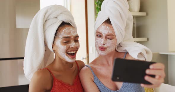 Diverse happy female friends wearing towels on heads and cleansing masks taking selfie at home alt