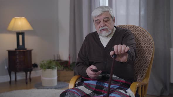 Portrait of Senior Caucasian Man with Walking Cane Switching Channels with Remote Control. Elderly alt