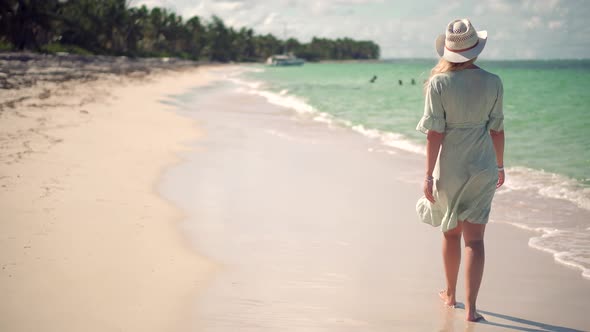 Woman In Hat Walks Along Beach On Caribbean Coast. Woman Relaxing On Seychelles Flowing Dress. alt
