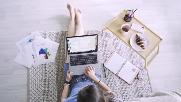 Young Woman with Laptop Sits on Floor Leaning on Bed By Tray