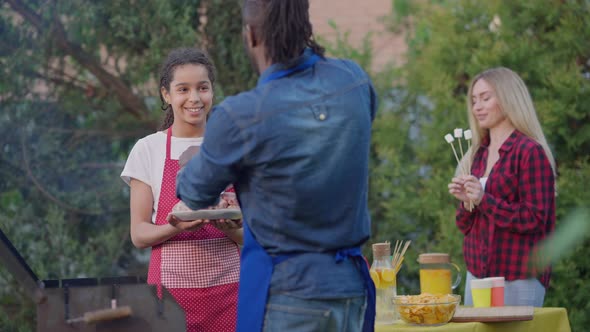 Teenage African American Girl Holding Plate with Raw Meat As Man Putting Burger Patties in Barbecue alt
