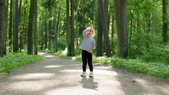 Cheerful Child in Striped Tshirt Waves Her Hand at Camera on Summer Sunny Day in Park Among Tall alt