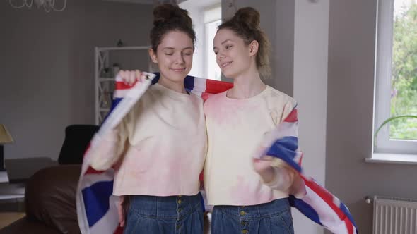 Medium Shot Smiling Adult Twin Sisters Wrapping in British Flag Hugging Looking at Camera alt