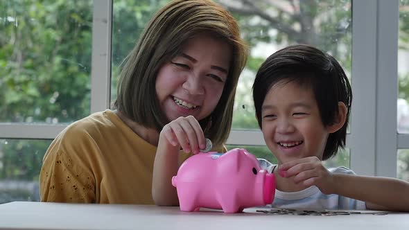 Cute Asian Child And Mother Putting A Coin In To A Piggy Bank alt