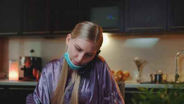 Pretty Business Woman in Pajama and with Medical Mask Speaking By Smartphone and Making Notes at alt