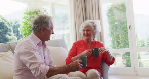 Happy caucasian senior couple sitting in sunny living room talking and drinking cups of coffee alt