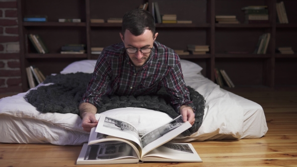 Young Man Lies on a Bed and Reads a Book at Home