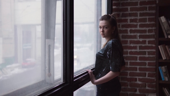 Young Woman with a Sad Look Is Standing By the Window alt