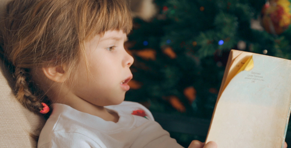 Small Girl Reading a Book in Front of Christmas Tree