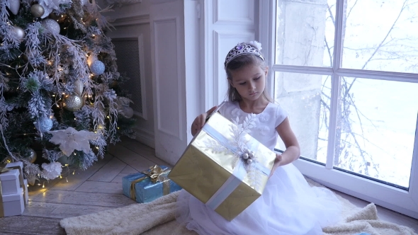 Little Adorable Girl Sitting Near the Christmas Tree and Looks at Christmas Gift