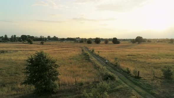 Aerial Footage of Young Woman Riding Bike Through Fields at Summer Sunset alt