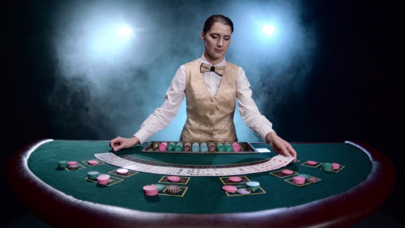 Girl Croupier at a Casino Shuffles Cards on Black Smoky Background with Spotlights. alt