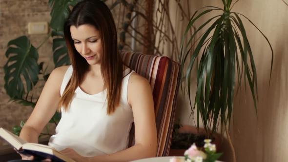 Young Beautiful Girl Reading a Book Sitting in a Chair. Attractive Woman Smiling While Reading