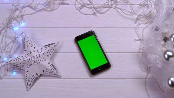 Mobile Phone Lies on a White Table, Next To Christmas Toys and Garlands. Top View alt