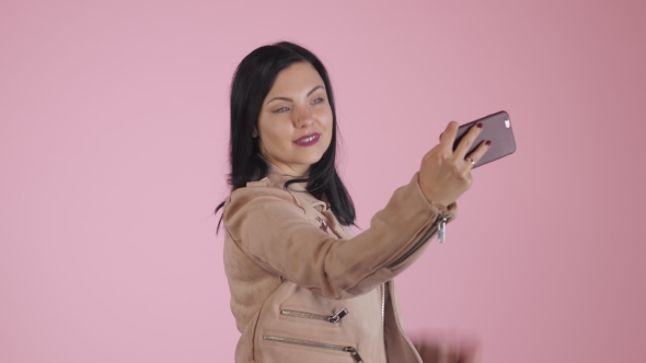 Smiling Happy Brunette Woman in Pink Jacket Making Selfie on Smartphone Over Colorful Background. alt