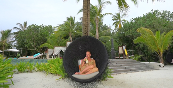 Girl Sitting in Luxurious Hanging Chair on Tropical Island Sipping a Cocktail alt