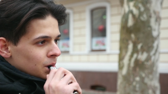 A Young Man Stands Outdoors and Smokes an E-cigarette in Winter