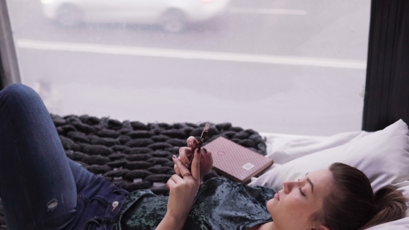 Young Woman Using Smartphone on Bed By Window Overlooking City Street